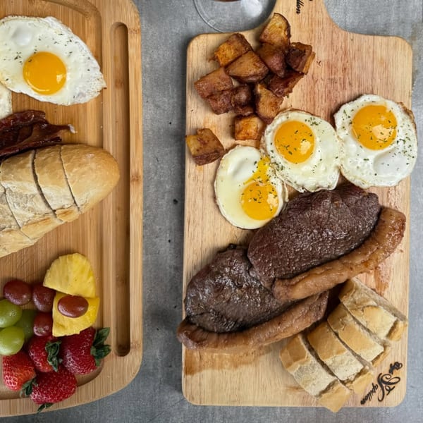 Two wooden boards with breakfast items: steak, eggs, potatoes, bread, bacon, and fruit.