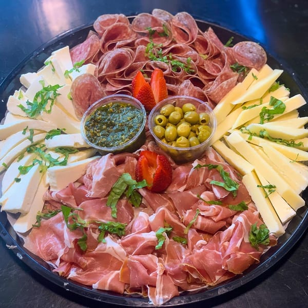 A large platter filled with various cold cuts, cheeses, olives, pesto, and strawberries.