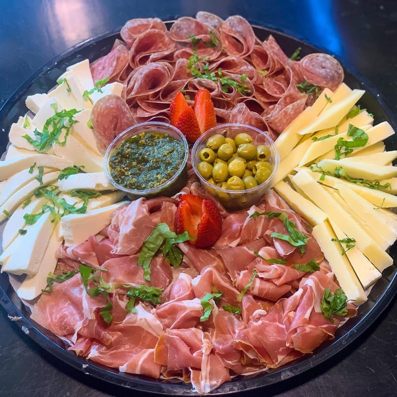 A large platter filled with various cold cuts, cheeses, olives, pesto, and strawberries.